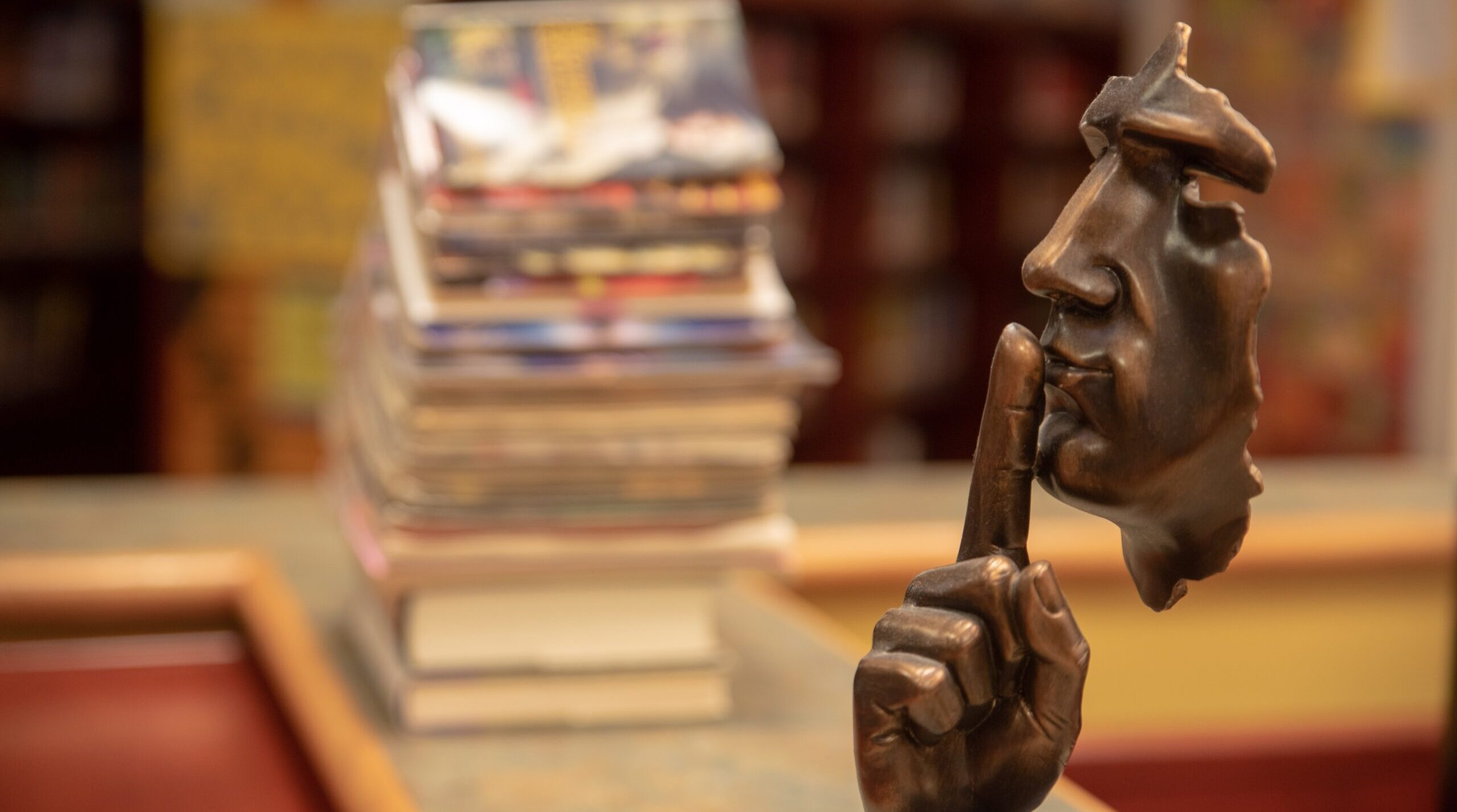 A small statue of a shushing finger in front of a face rests on a shelf. In the background, stacks of books. What information do we lose by removing sound from our texts?