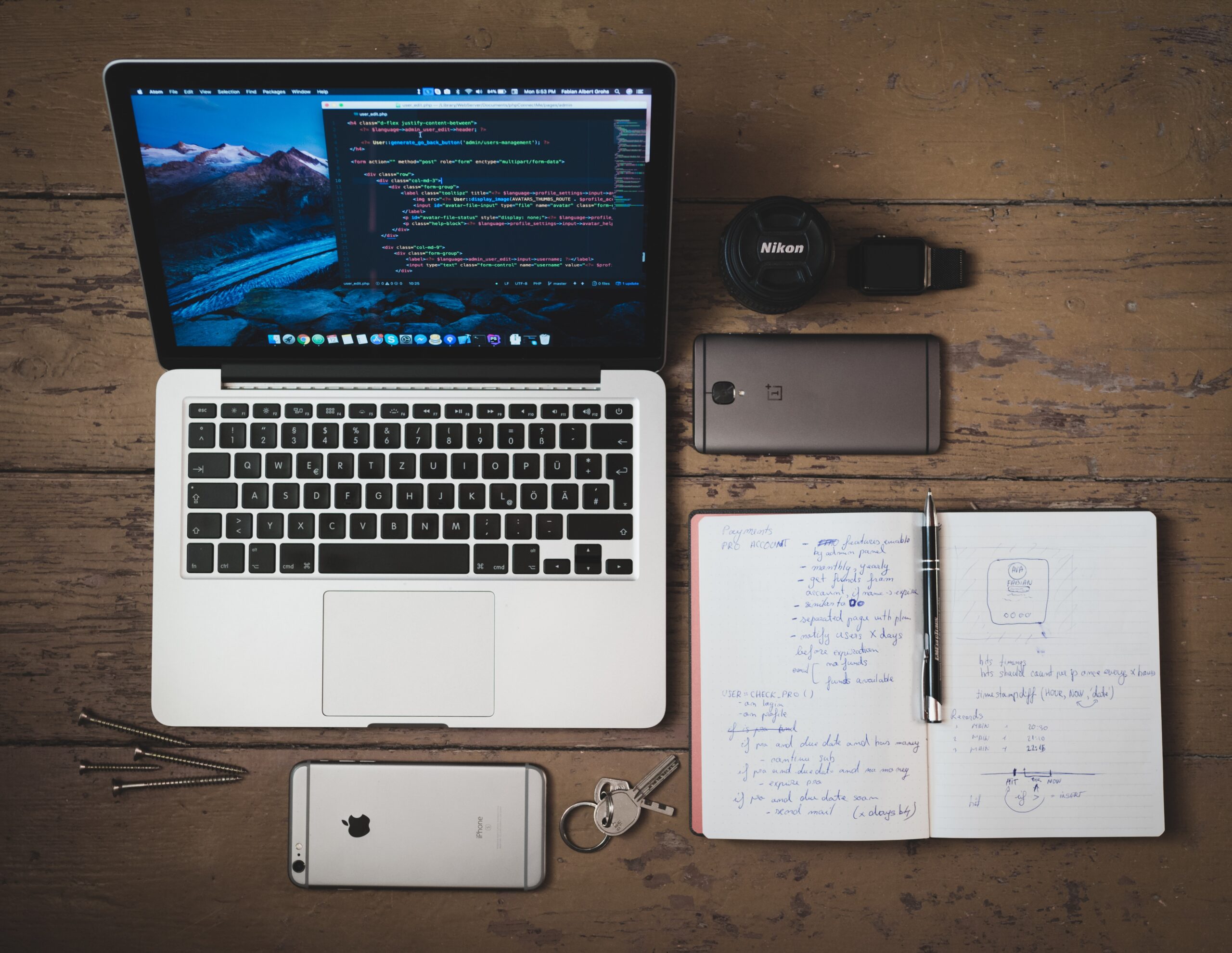 A laptop with coding on the screen sits on a desk next to two smartphones, a pair of headphones, an open notebook with writing inside, a pair of keys, and four screws.