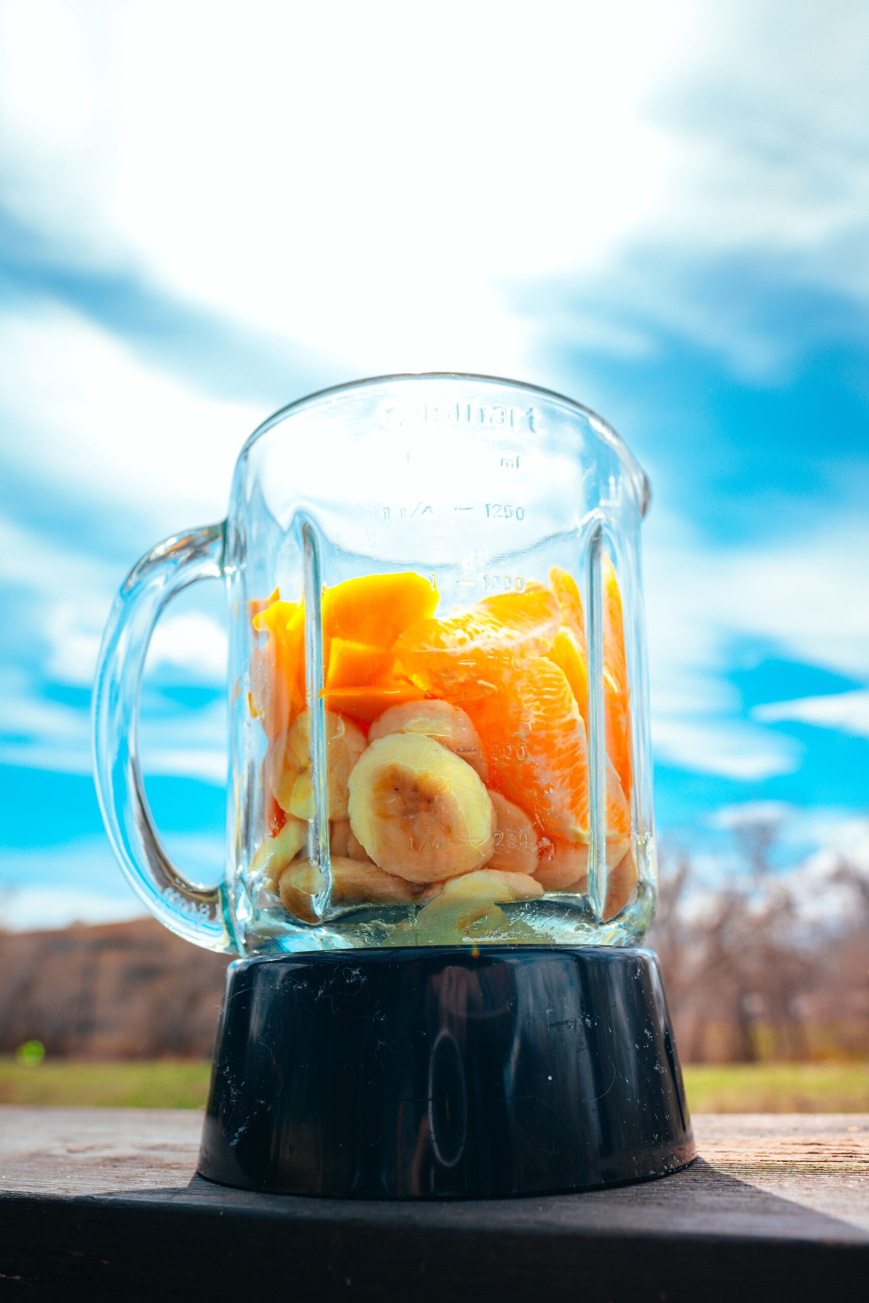 A photo of a blender filled with sliced banana and oranges taken from an upwards angle. The blender lacks a lid, and is placed outside in front of a bright blue sky.