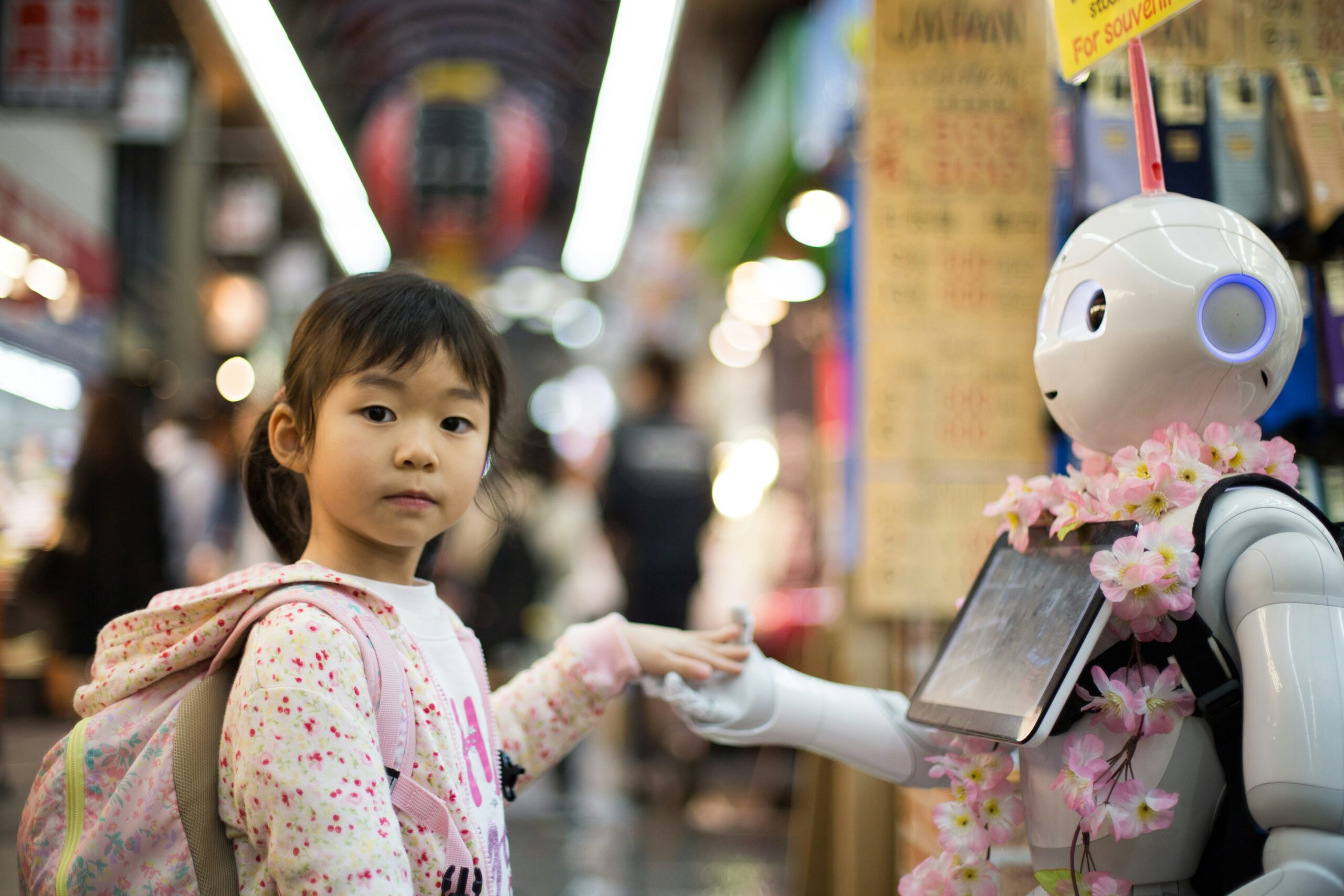 A young Asian girl with a pink patterned hoodie holds hands with a robot. It is white, made of smooth plastic with rounded features. It has a lei of pink flowers around its neck. Both look towards the camera.