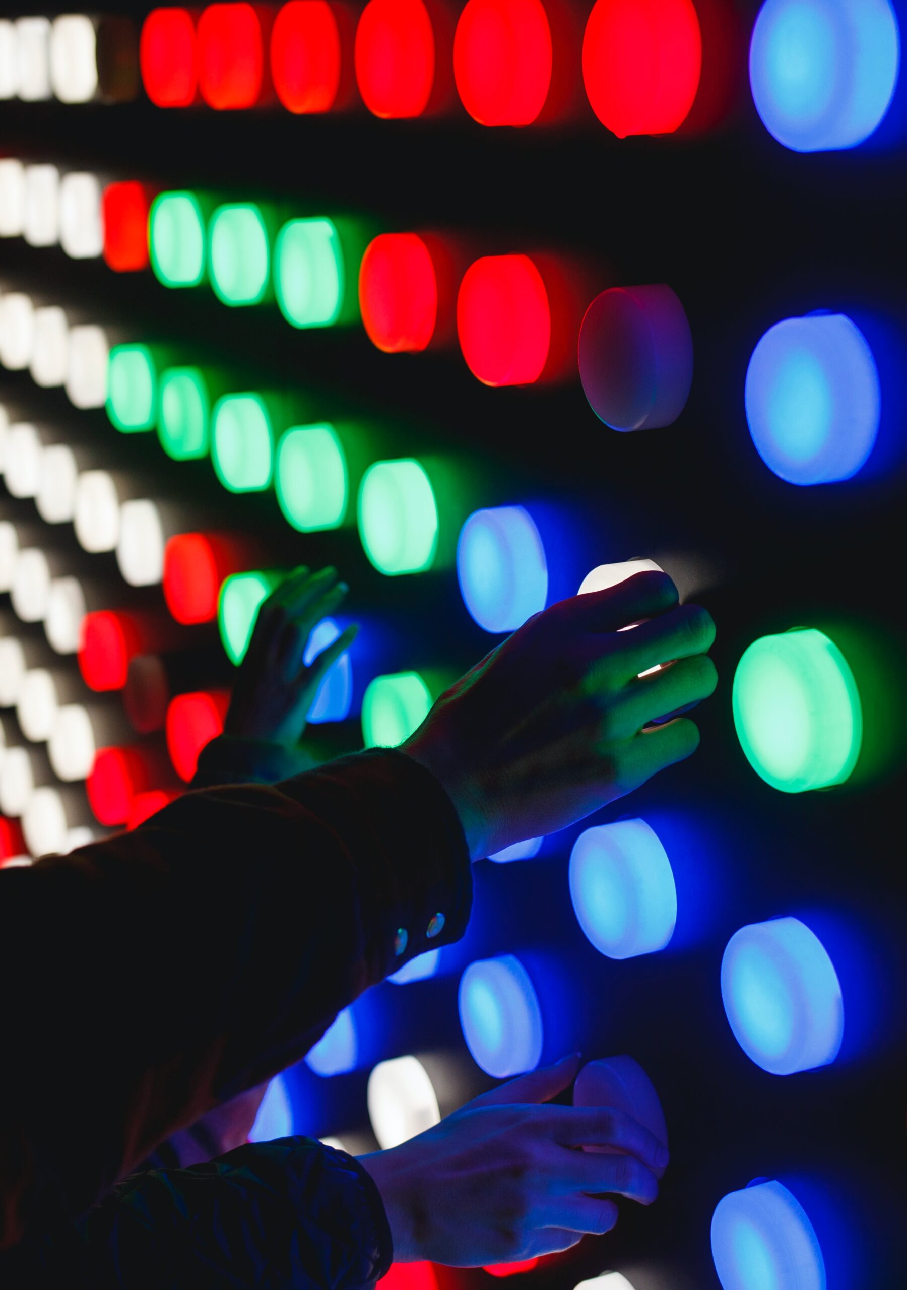 A dark wall of lit up colored buttons. To the far left, the buttons are white, and towards the right, they become red, green, and blue. A few hands reach out to touch the buttons.