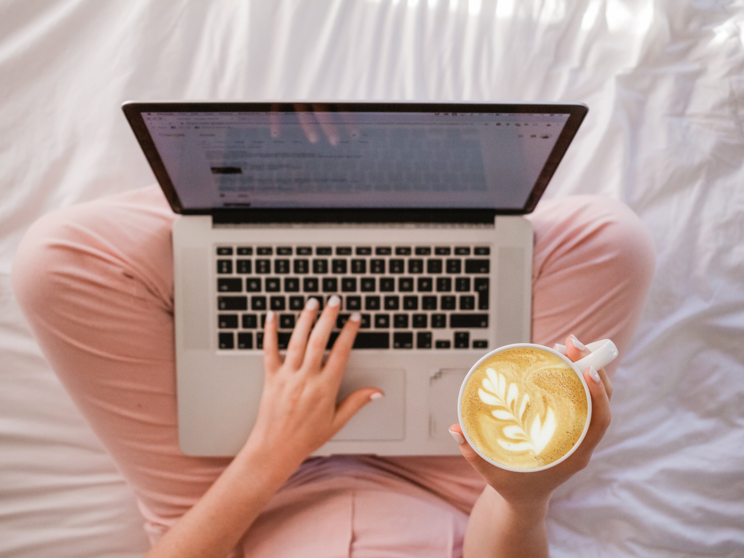 An overhead view of a laptop in the lap of a woman wearing pink lounge pants. She holds a teacup with latte art in one hand.