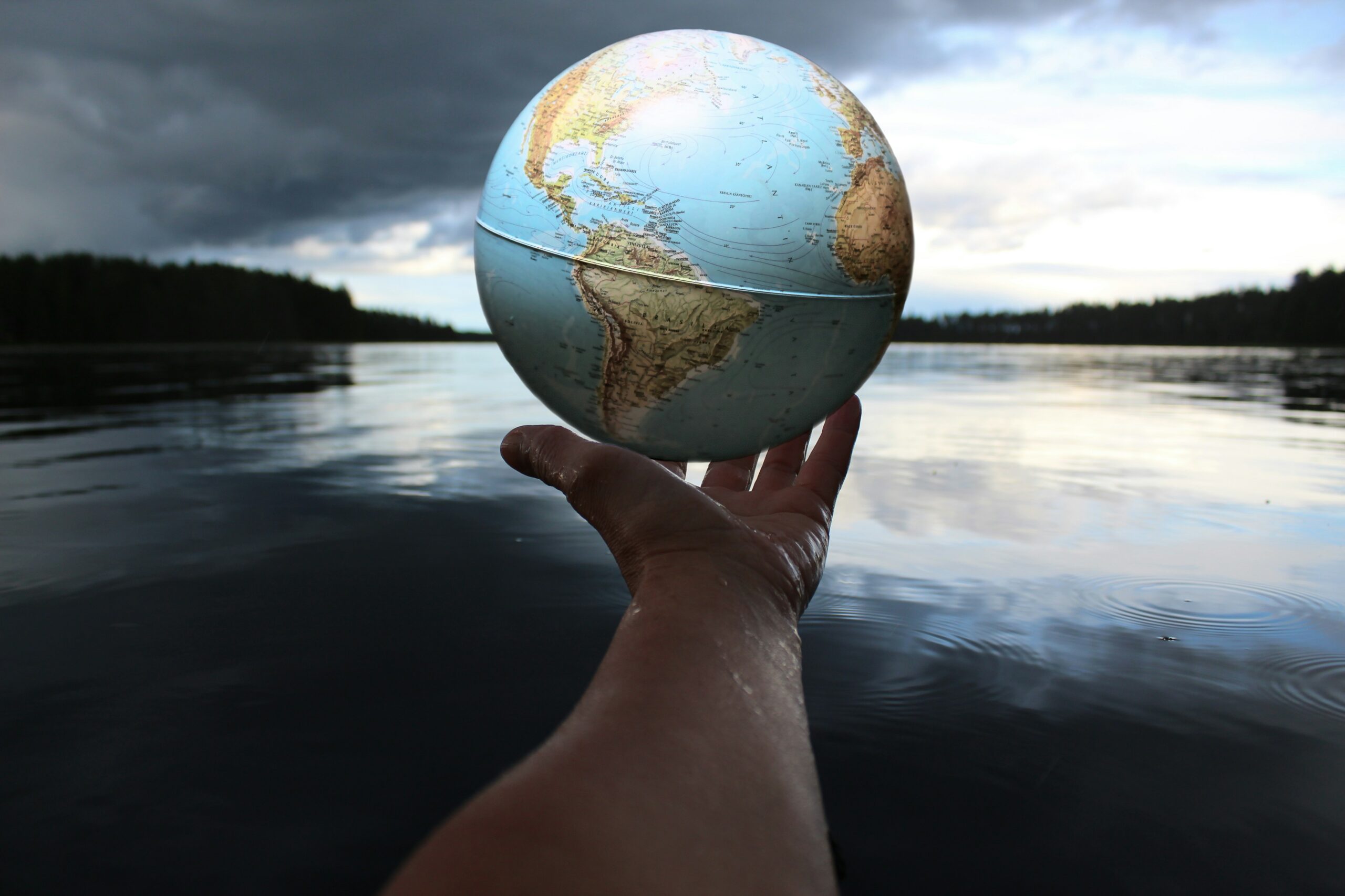 This is an image of a person holding a globe map while they are surrounded by nature and a lake in front of them. The entire image represents as both digital and visual rhetoric, but more digital.