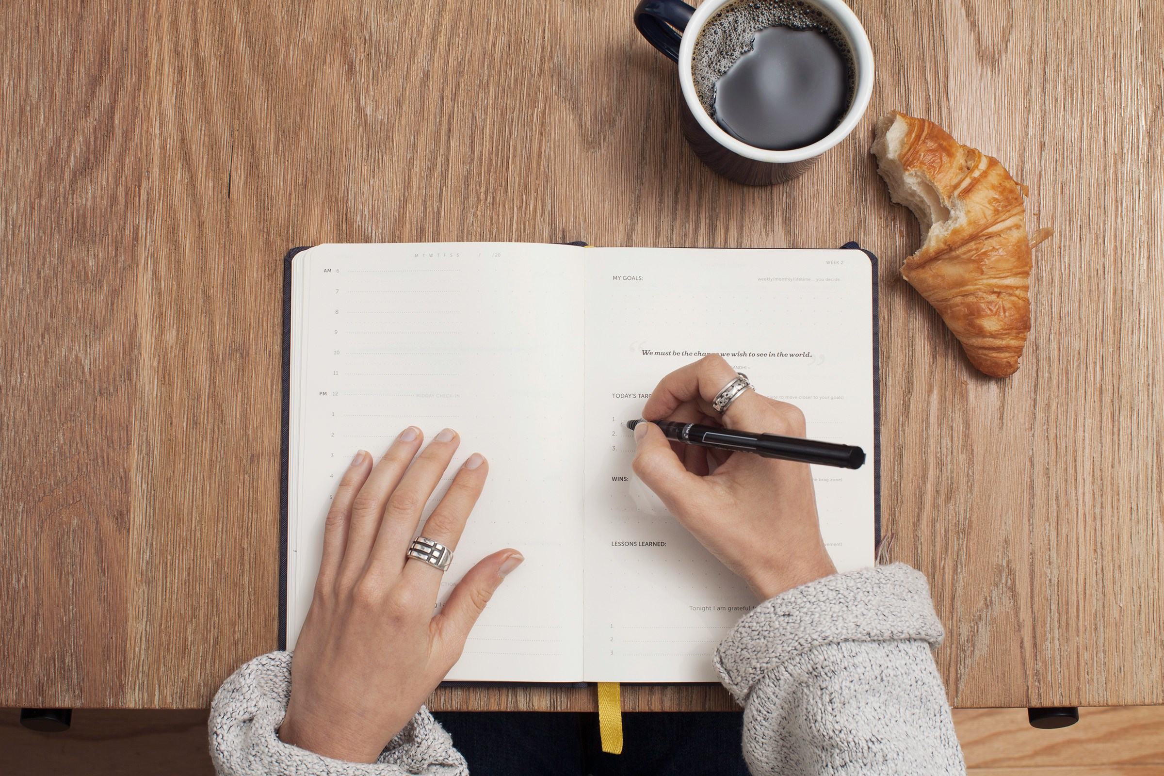 Person writing in a notebook with a bitten croissant and black coffee sitting in front of the notebook