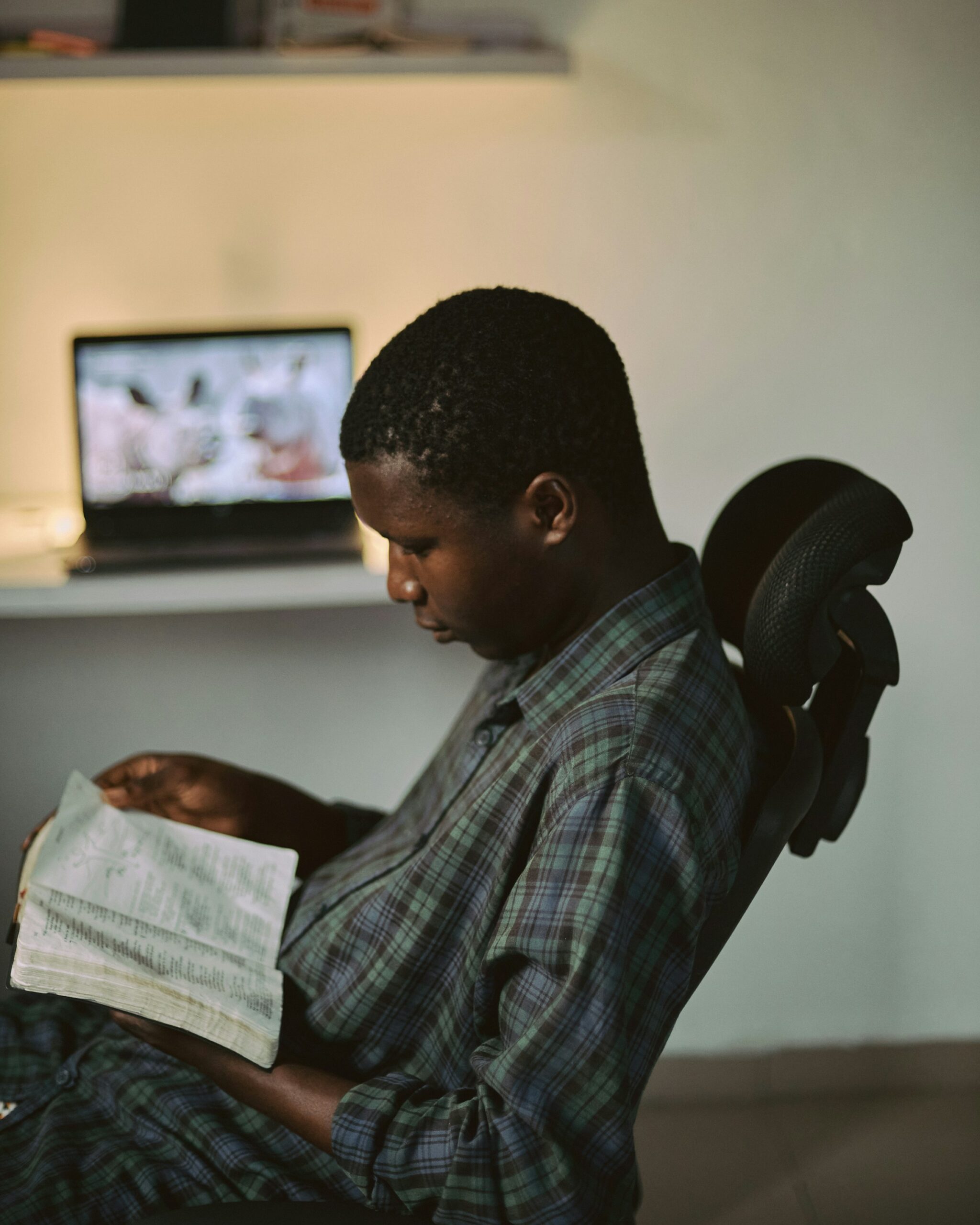 This is an image of a man reading a book (form of old media) while the computer in the back of the room is just sitting there.