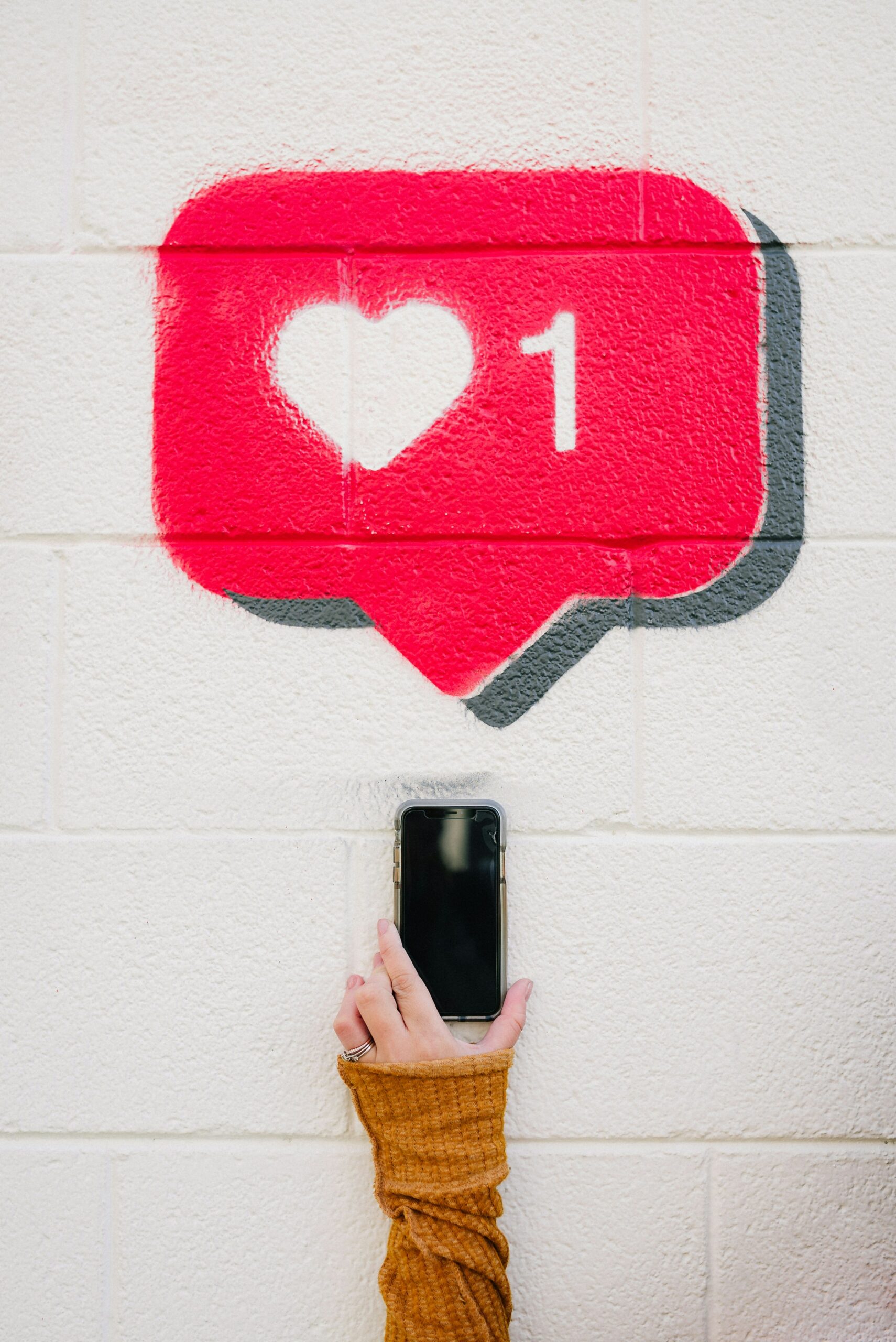 This is an image of large heart symbol with a "1" next to it that's in a text bubble. Underneath the text bubble is a phone and someone holding it. This image represents that comments and likes demonstrate interactivity.
