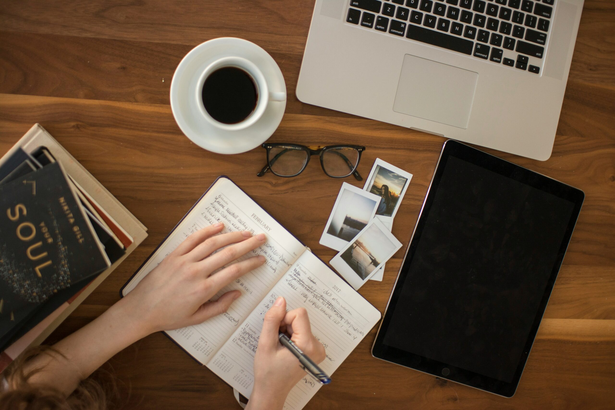 An image of books, a tablet, the keyboard of a laptop, a pair of glasses, a cup of coffee, photographs, and an open notebook scattered on a hard wood table. One hand is resenting on the left side of the notebook while the other is writing with a ballpoint pen on the right side of the book. This person has a variety of digital and analog devices on their table!