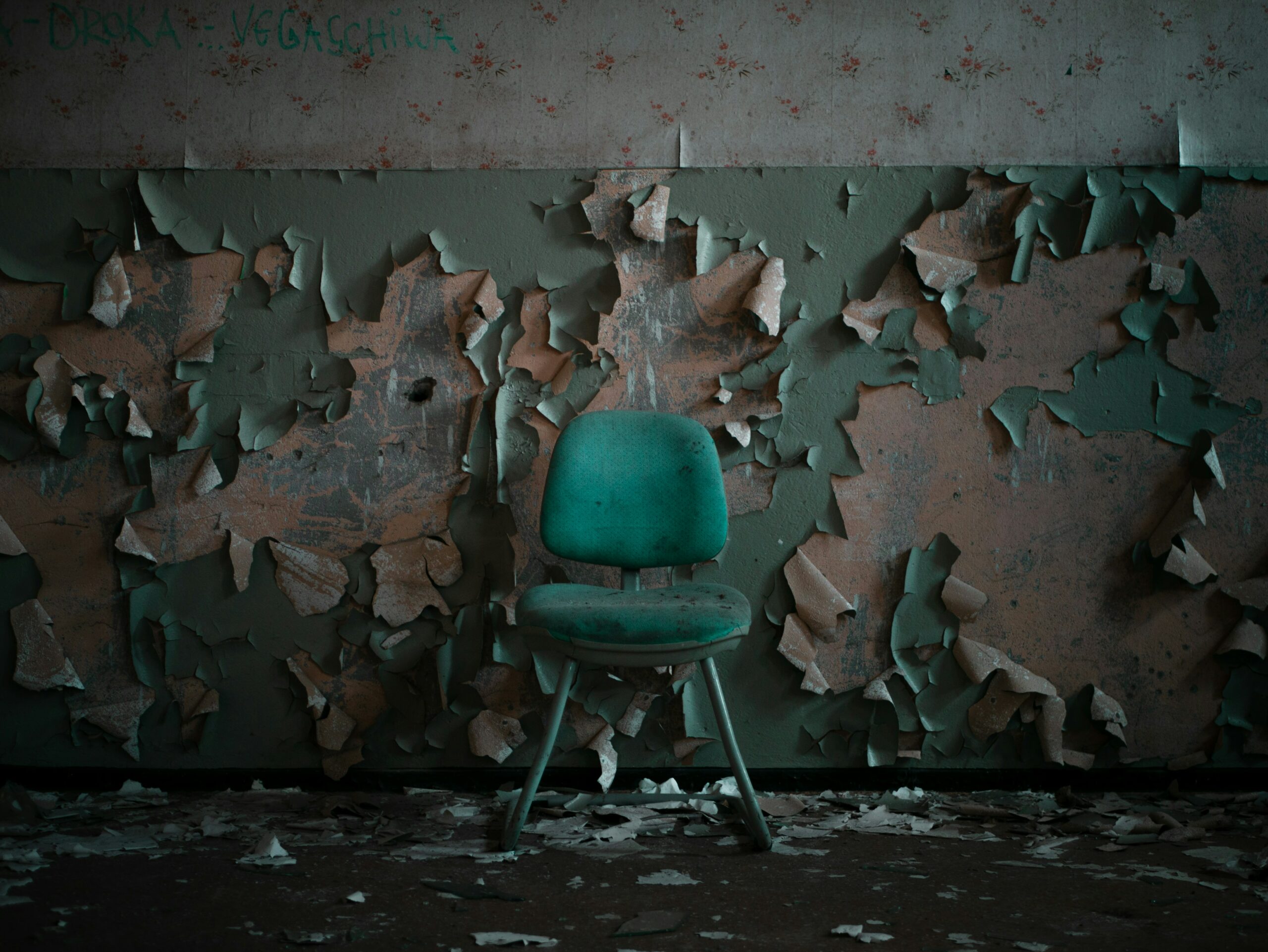 In the middle of a dark room, there's a dirty teal chair. The paint on the wall behind it is peeling, and the floor is a mess.