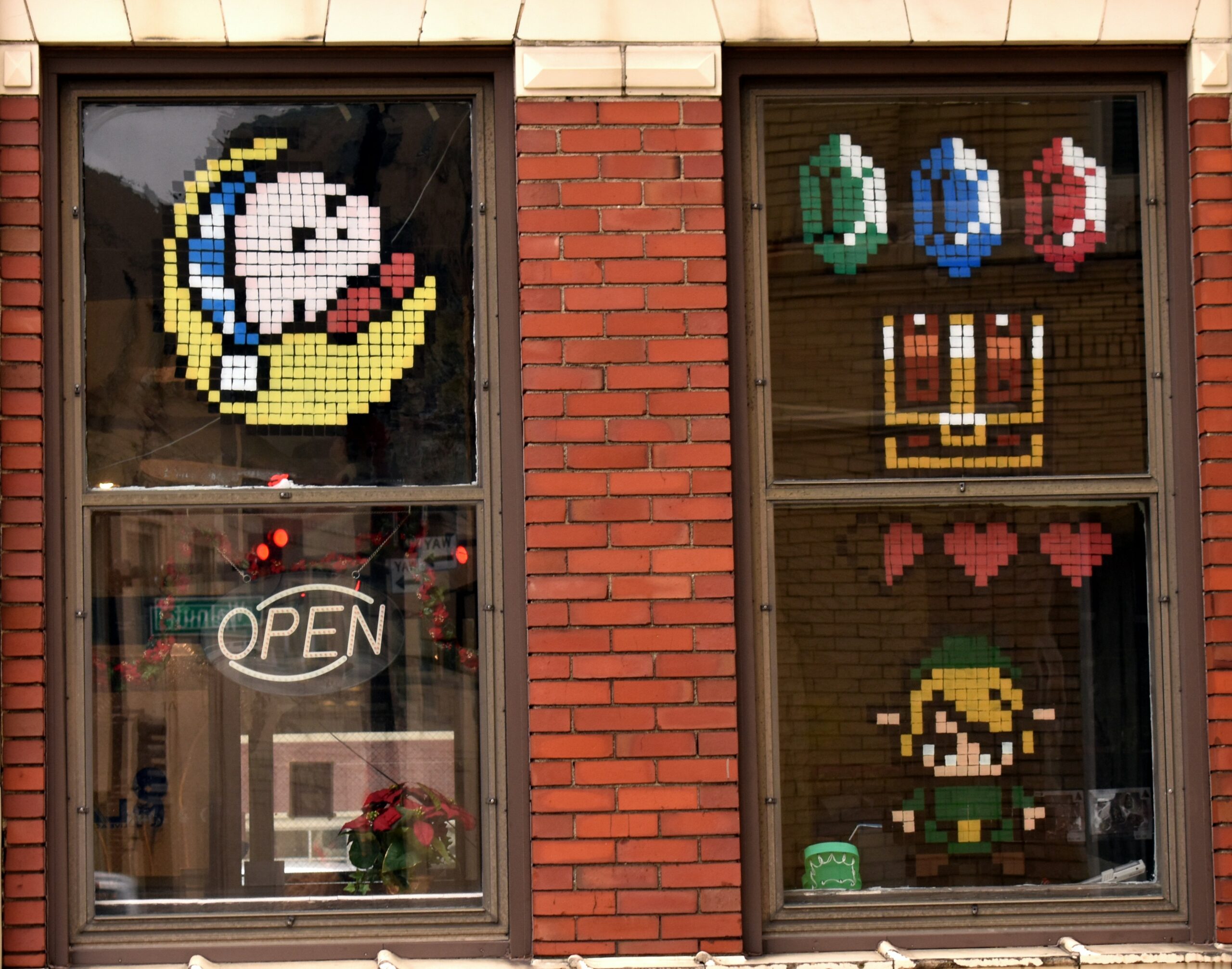 The outside of a brick wall store. It has two windows: One has pixel art of Kirby sleeping atop a moon plastered on it. The other has pixel art of the rupees, chest, and hearts from Legends of Zelda. There's a mini Link, too.