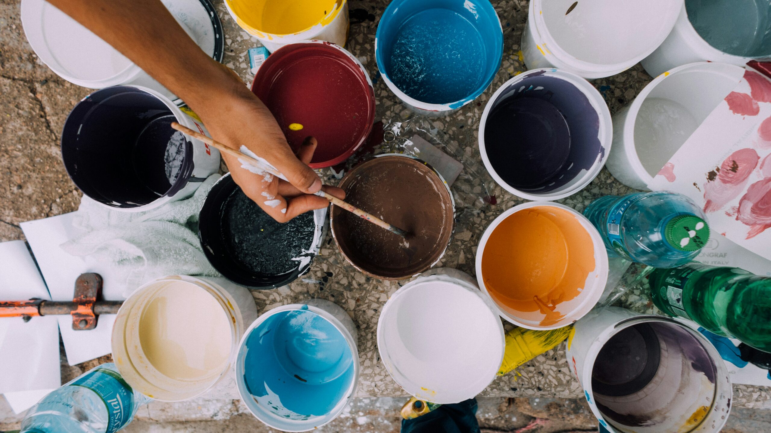 An image of many open paint cans. A single hand, stained with white paint, dips the tip of a paintbrush in a can of brown paint. A user interface utilizes the art of design to be a convenient point of contact between a computer and person.