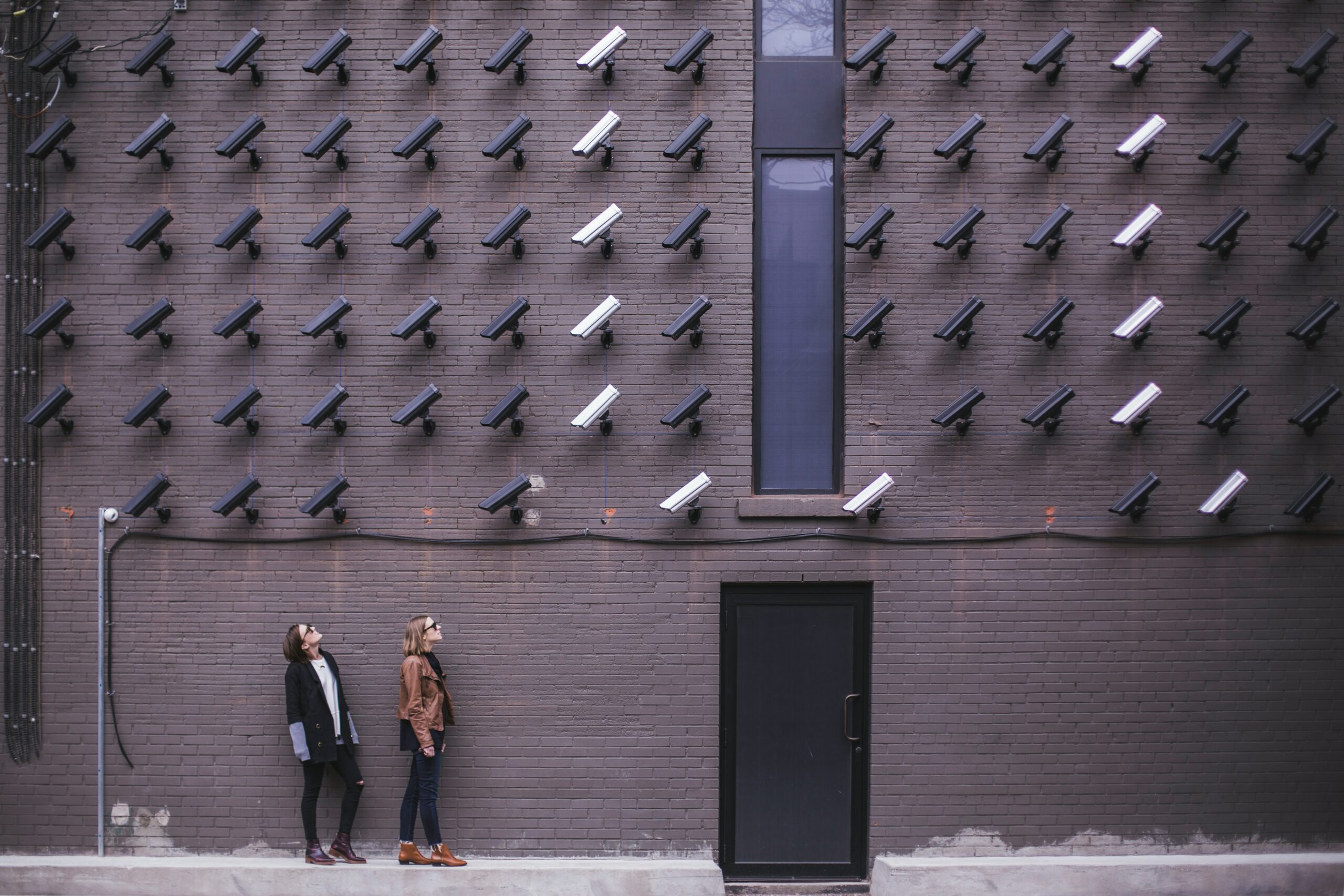Two women stand against a brick wall, both are looking up at many surveillance cameras that are all pointed in the women's direction.