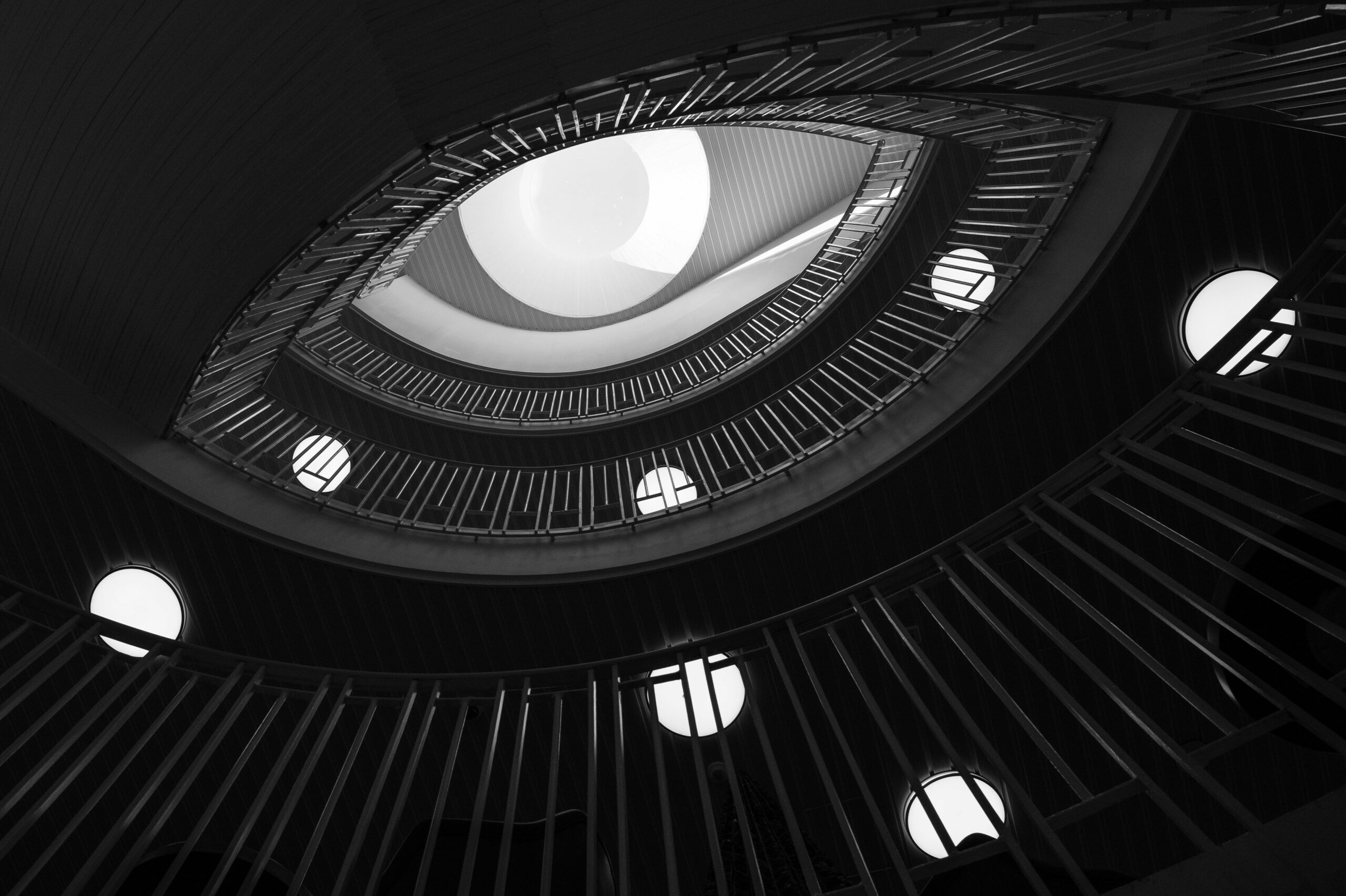 A grayscale photo of a stairwell. The railings are illuminated mainly by a large, white ceiling lamp on the top floor. The shape of the railings, combined with the ceiling and it's lamp resemble an eye glaring down at the viewer.