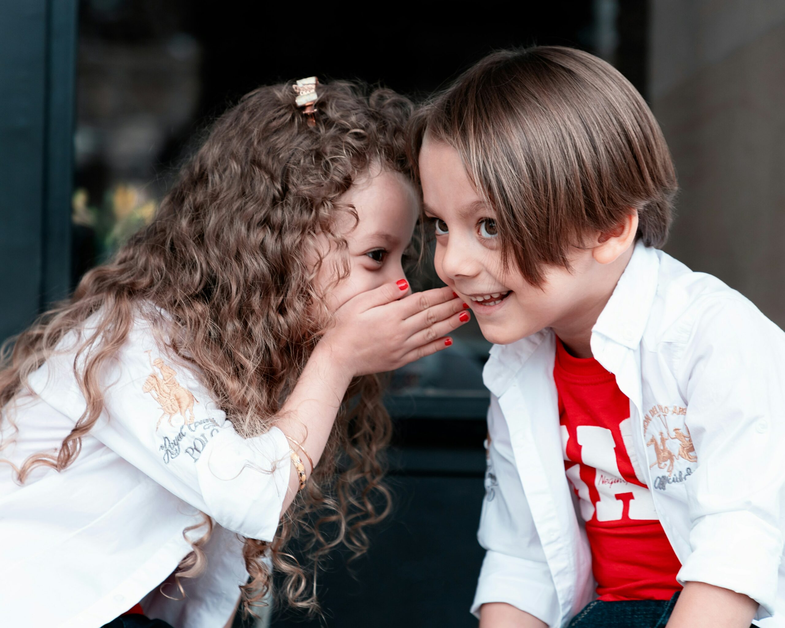 A little girl whispers to a little boy with her hand up to his ear.
