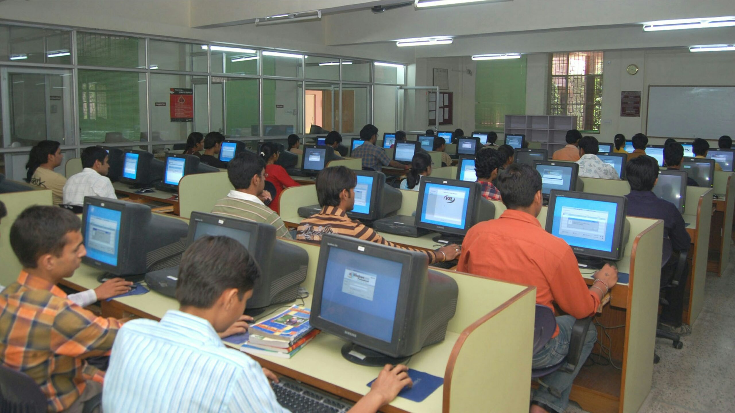The image shows a classroom of students in a computer lab. The whole room is full of computers popular in the early 2000s with a student sat in front of every one.